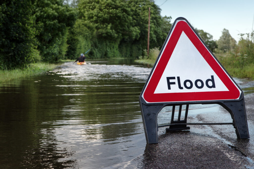 Flooded road with a flood warning sign and a person rowing on the water in the background.